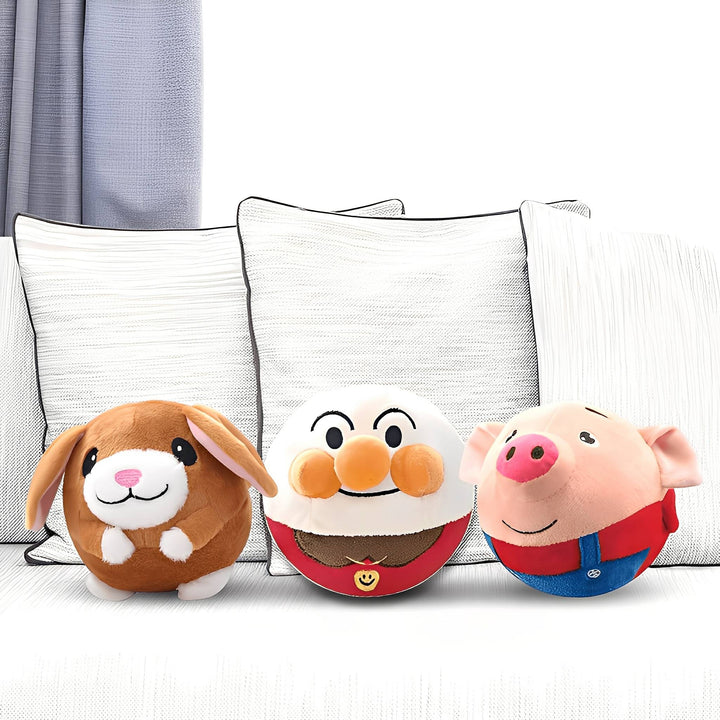 Drei runde Plüschbälle als Hase, Gesicht mit orangefarbener Nase und Schwein nebeneinander vor weißen Sofakissen. 1